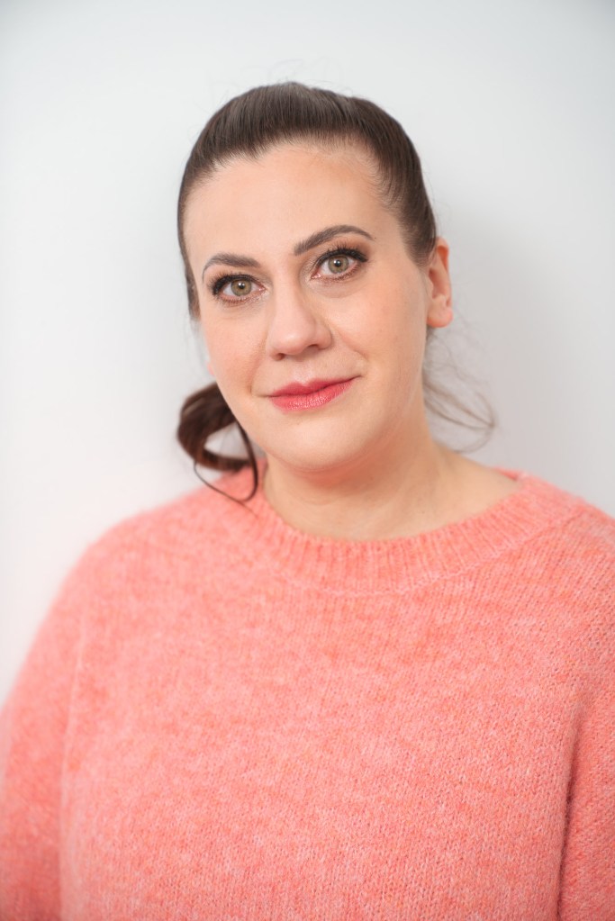 A woman with dark hair tied back, wearing a pink sweater, standing against a white background and looking directly at the camera.