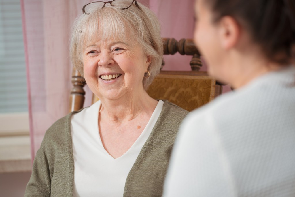 An older woman with gray hair and glasses smiling while seated in a chair, engaged in conversation with another person.