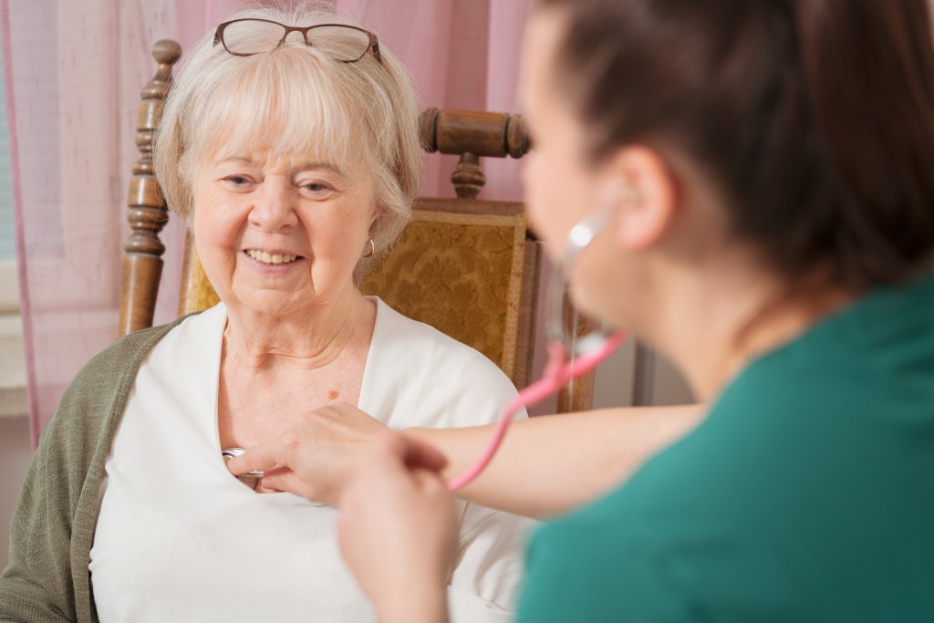 A healthcare professional using a stethoscope to check the heartbeat of an elderly woman sitting in a chair, smiling.