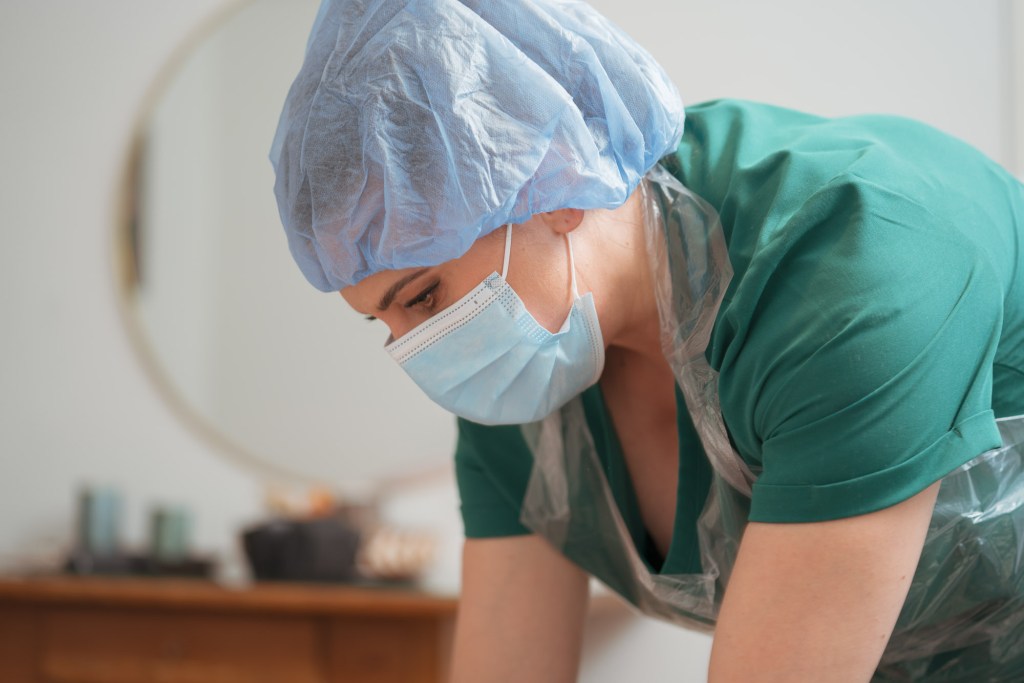 A healthcare professional wearing a blue surgical cap and mask, looking down intently while working, dressed in a green scrubs top and a protective plastic apron.