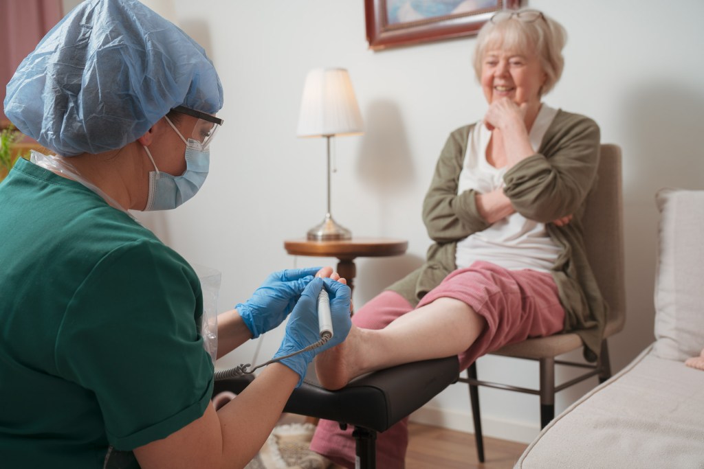 A healthcare professional in protective gear is performing a foot treatment on an elderly woman, who looks relaxed and happy while sitting on a couch in a well-lit room.